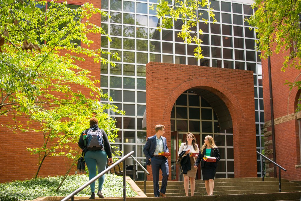 Entrance to a brick building with large windows featuring a grid of glass panes. The doorway is framed by a red brick arch and leads to a set of double doors. The entrance is accessed by a semi-circular set of stairs with black handrails.